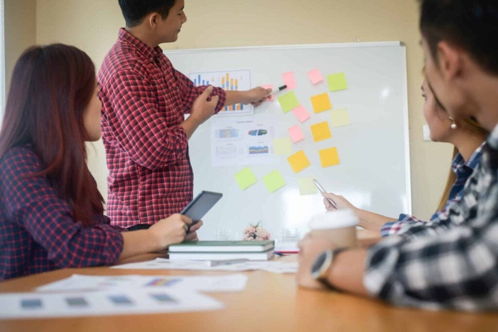 Young man presenting data on whiteboard during group meeting with sticky notes.